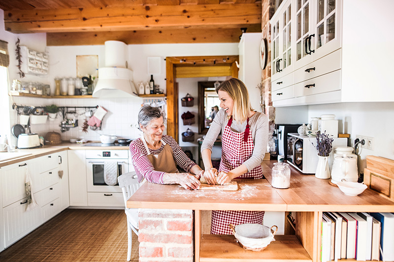 elderly-grandmother-adult-granddaughter-cooking inkitchen Smiling grandmother and granddaughter baking together in a cozy kitchen, rolling dough on a wooden counter.