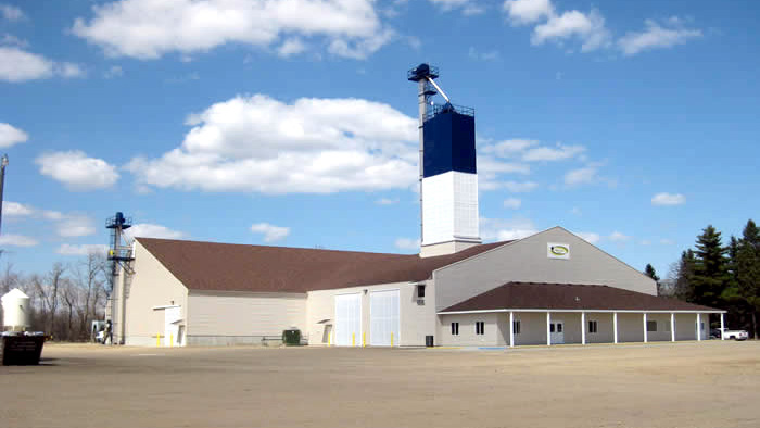 Large agricultural building with tan siding, brown roof, and a tall blue and white grain leg tower, set against a blue sky with scattered clouds.