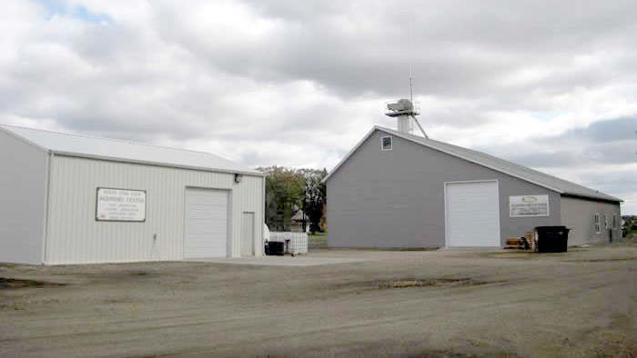 Two large agricultural buildings with metal siding and overhead doors, one white and one gray, standing on a dirt lot under a cloudy sky.