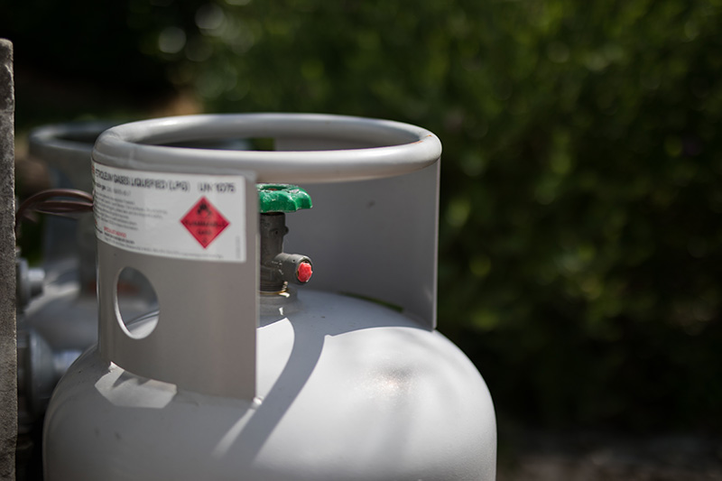 propane-tanks Close-up of a propane tank cylinder with a green valve and safety warning label, set outdoors with greenery in the background.