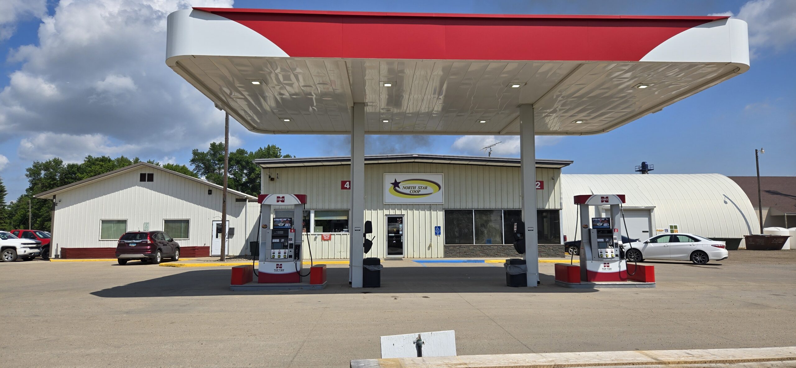 North Star Coop gas station with two fuel pumps under a red and white canopy in front of a cream-colored building, with parked vehicles and a clear sky in the background.
