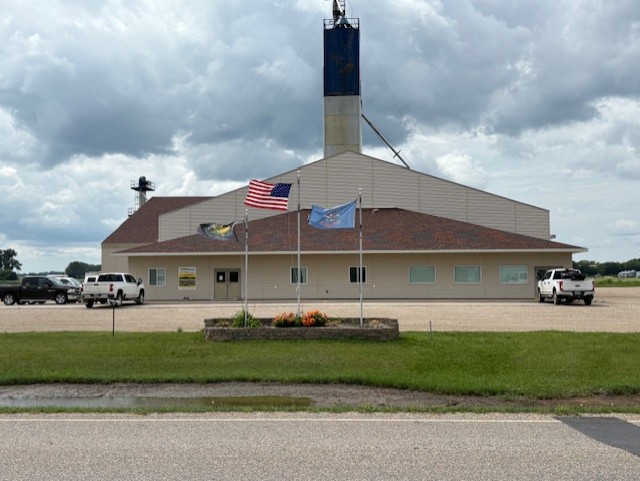 Large cream-colored agricultural building with a tall blue grain leg tower, three flags flying in front (including the U.S. flag), and several trucks parked around the lot under a cloudy sky.