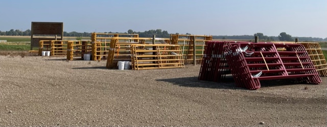 thumbnail_IMG_3482 Outdoor lot with stacks of metal livestock gates and panels in yellow and red, arranged on gravel with farmland in the background.