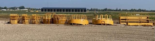 thumbnail_IMG_3483 Outdoor lot with rows of yellow livestock feed bunks and bale feeders lined up on gravel, with farmland and a large black-roofed building in the background.